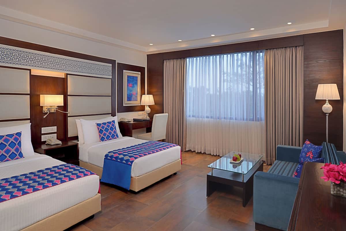 A hotel room with two beds, a sofa, a glass coffee table, and a desk with a lamp.