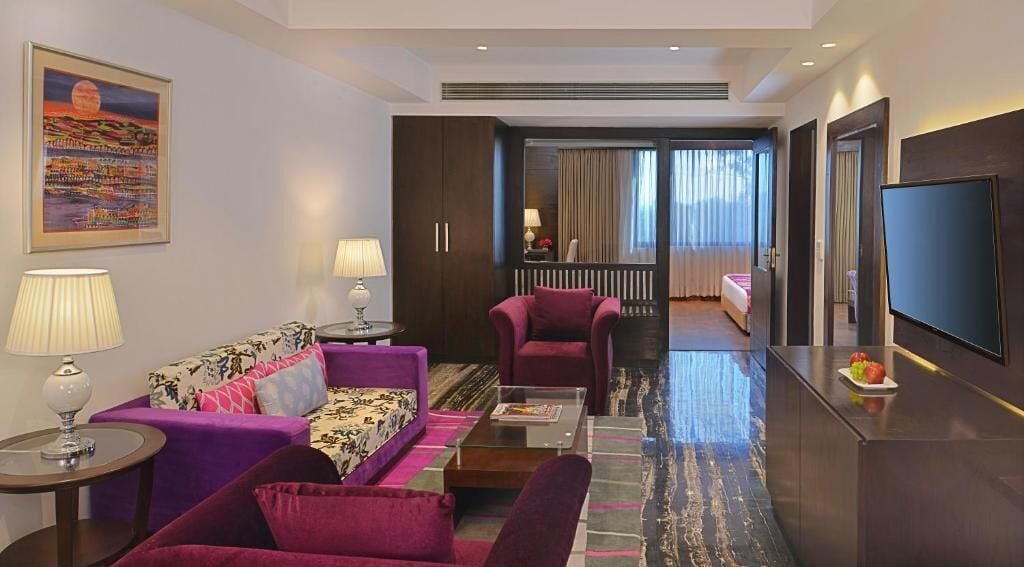 A hotel room with a sofa, armchair, and a bed. There is a TV on the wall and a window with curtains.