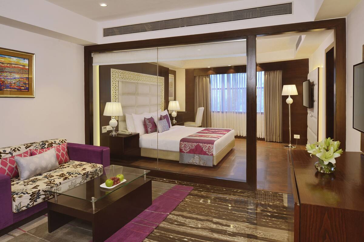 A hotel room with a bed, a sofa, a coffee table with fruit, a TV, and a painting on the wall.