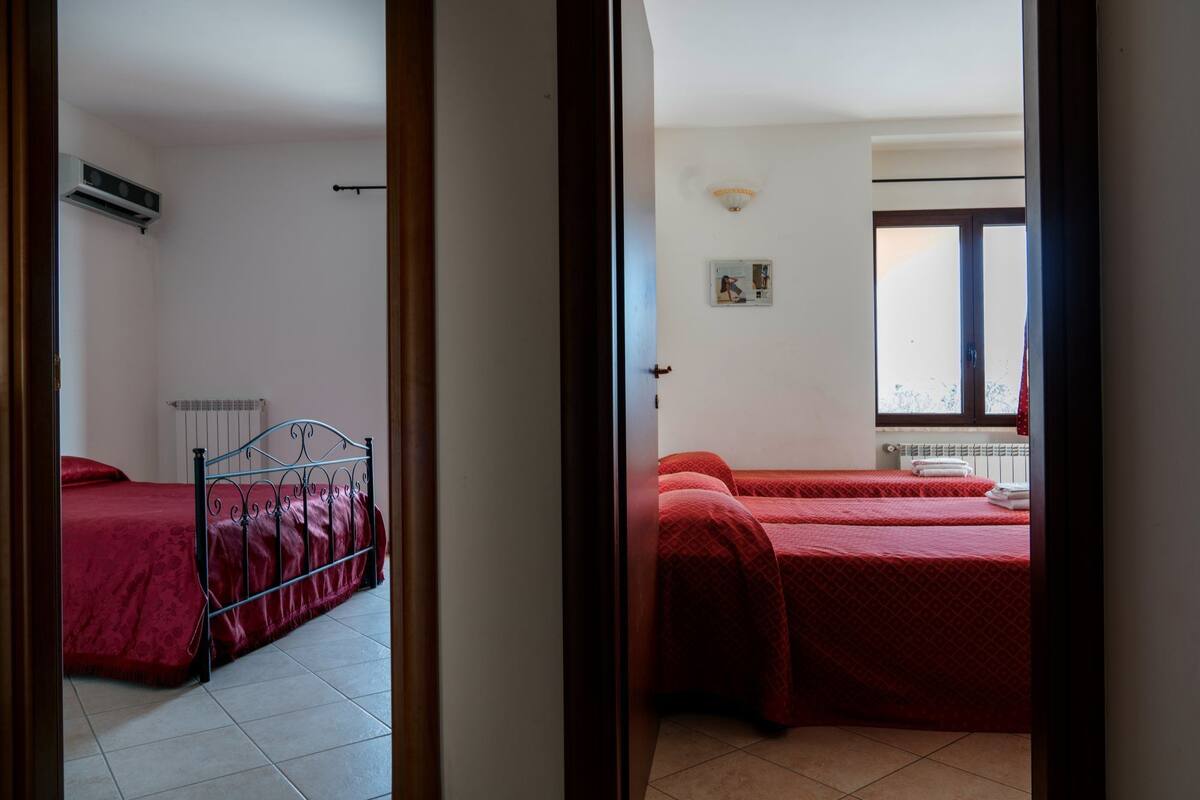 Habitación con dos camas, una con estructura metálica y ropa de cama roja, y otra con estructura de madera y ropa de cama roja. Hay un radiador debajo de la cama con estructura metálica, una ventana con vista al exterior y una lámpara fijada a la pared.