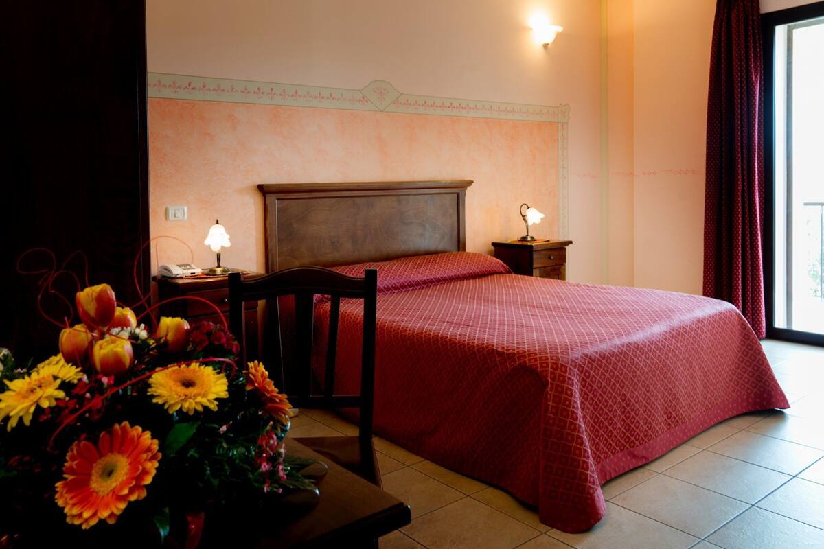 Habitación de hotel con una cama grande, dos mesitas de noche con lámparas, un cabecero de madera, un arreglo floral a la izquierda y vistas al exterior a través de una ventana.