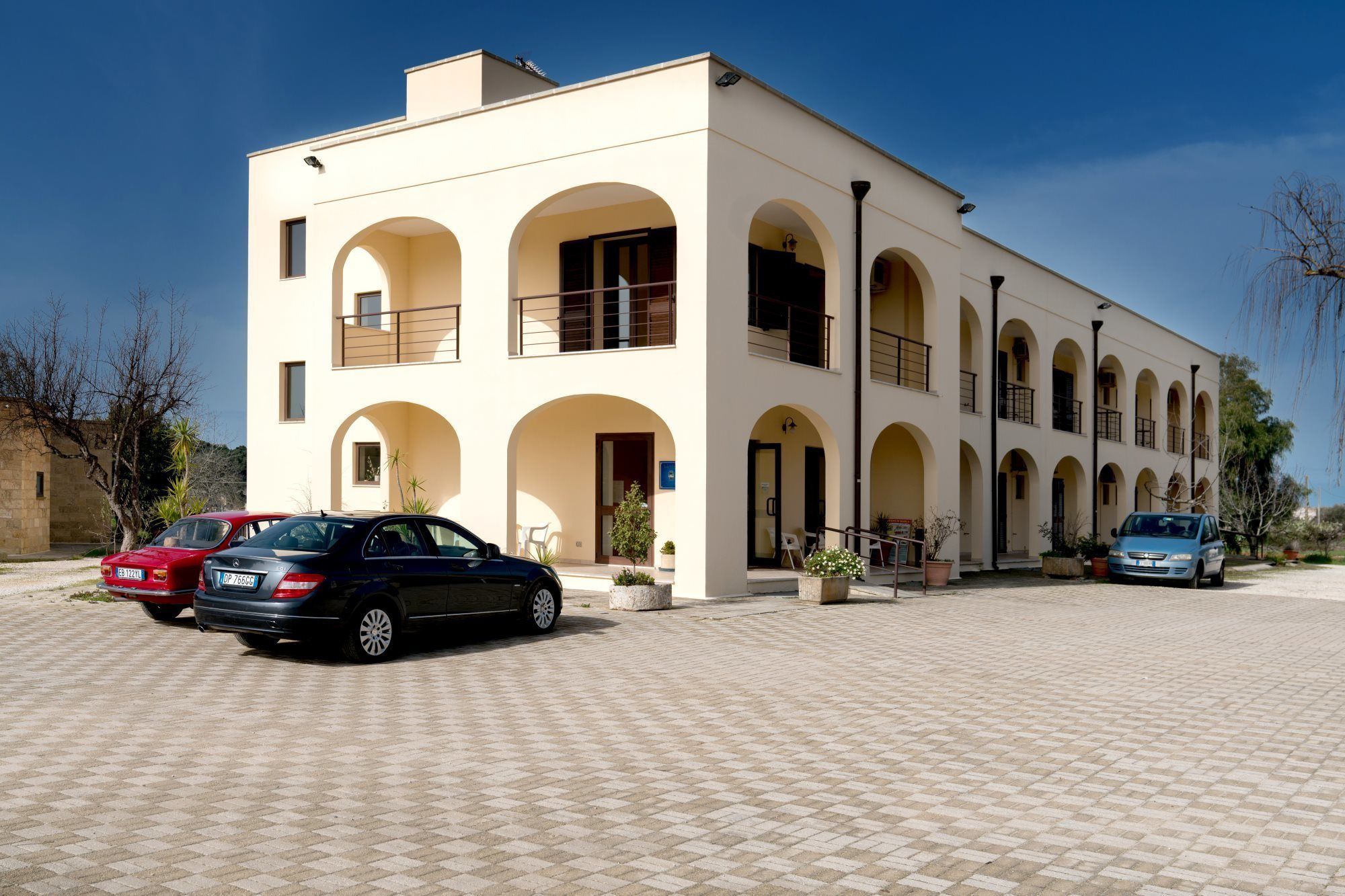 Ein modernes Gebäude mit weißer Fassade, mehreren Balkonen und einem gepflasterten Innenhof mit parkenden Autos.
