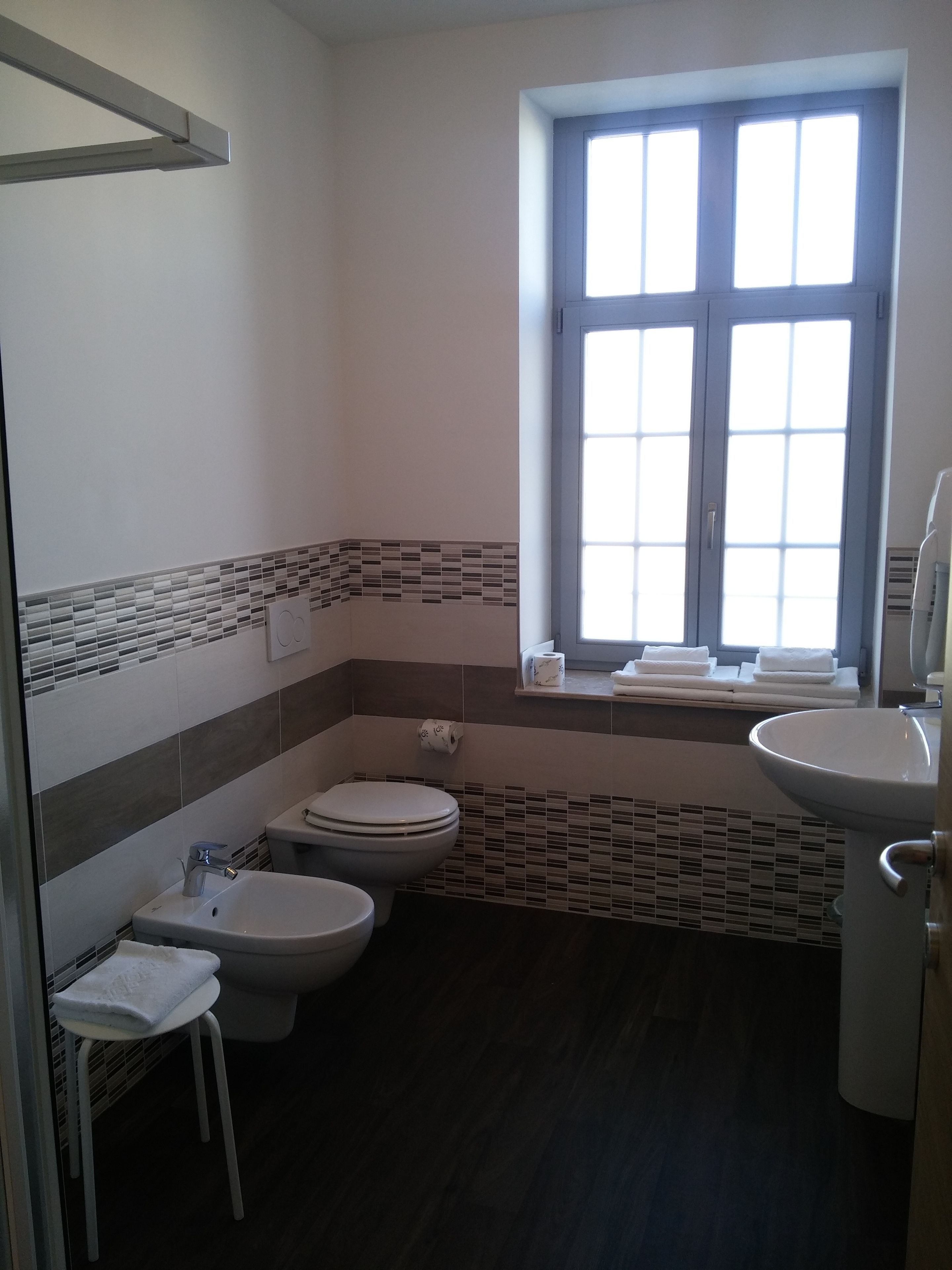 A bathroom with a toilet, bidet, and sink. There is a window with blinds and a towel rack.