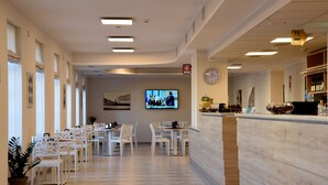Un vestíbulo de hotel con sillas y mesas blancas, un mostrador de recepción, un reloj y una pantalla de televisión.