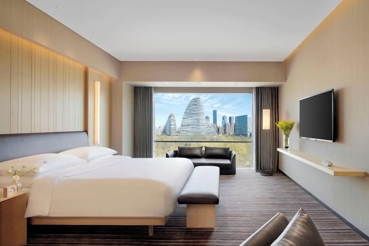A modern hotel room with a large bed, a sofa, a TV, and a city view.