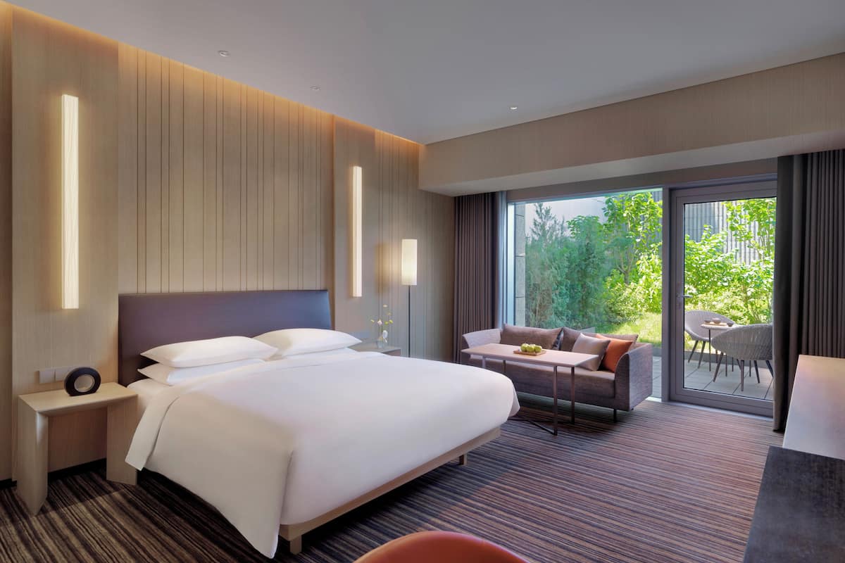 Habitación de hotel moderna con una cama grande, un sofá, una mesa de comedor y vistas a zonas verdes.