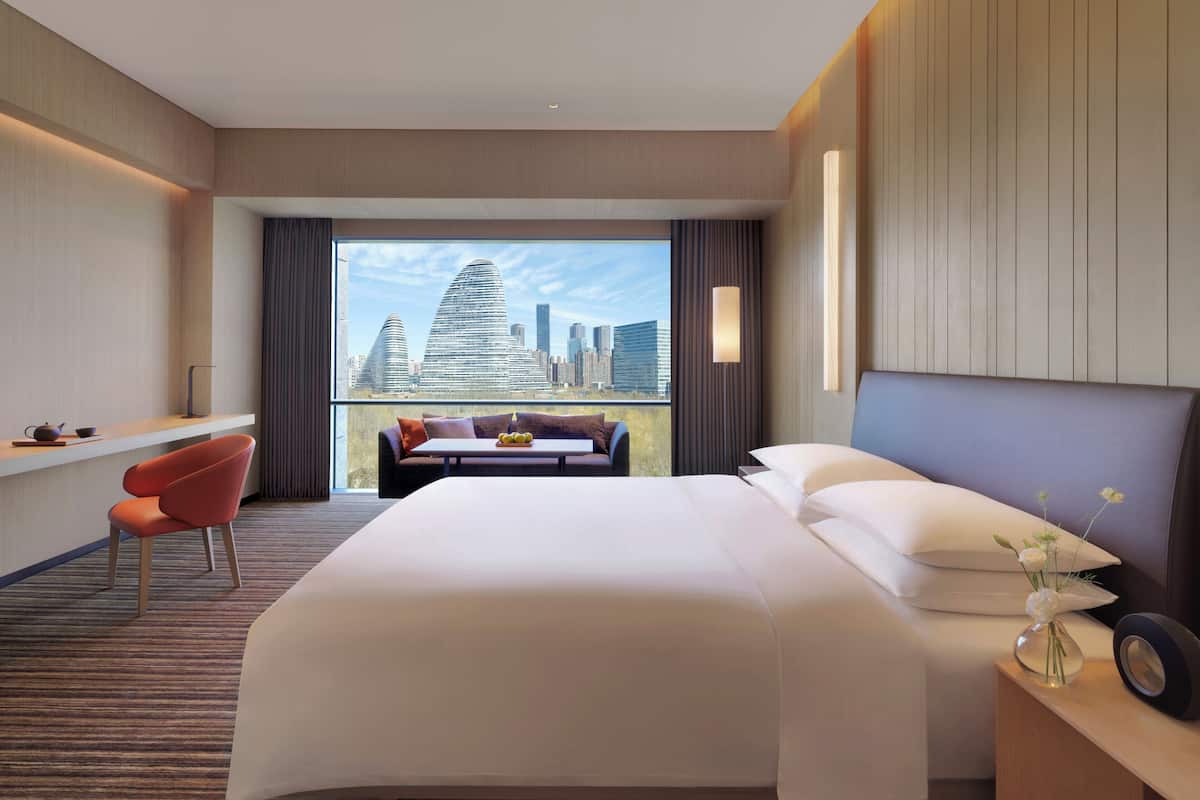 Habitación de hotel con una cama grande, zona de estar con vistas al horizonte de la ciudad, escritorio con silla y ventana con cortinas.