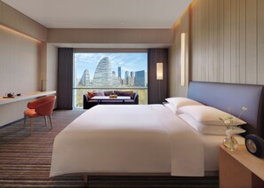 A hotel room with a large bed, a seating area with a view of the city skyline, a desk with a chair, and a window with curtains. - Hyatt Regency Beijing Wangjing (Beijing)