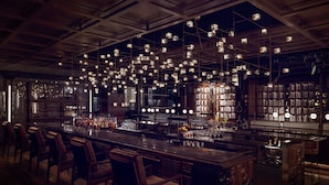 Zona de bar con sillas de madera, una barra alargada y luces colgantes.