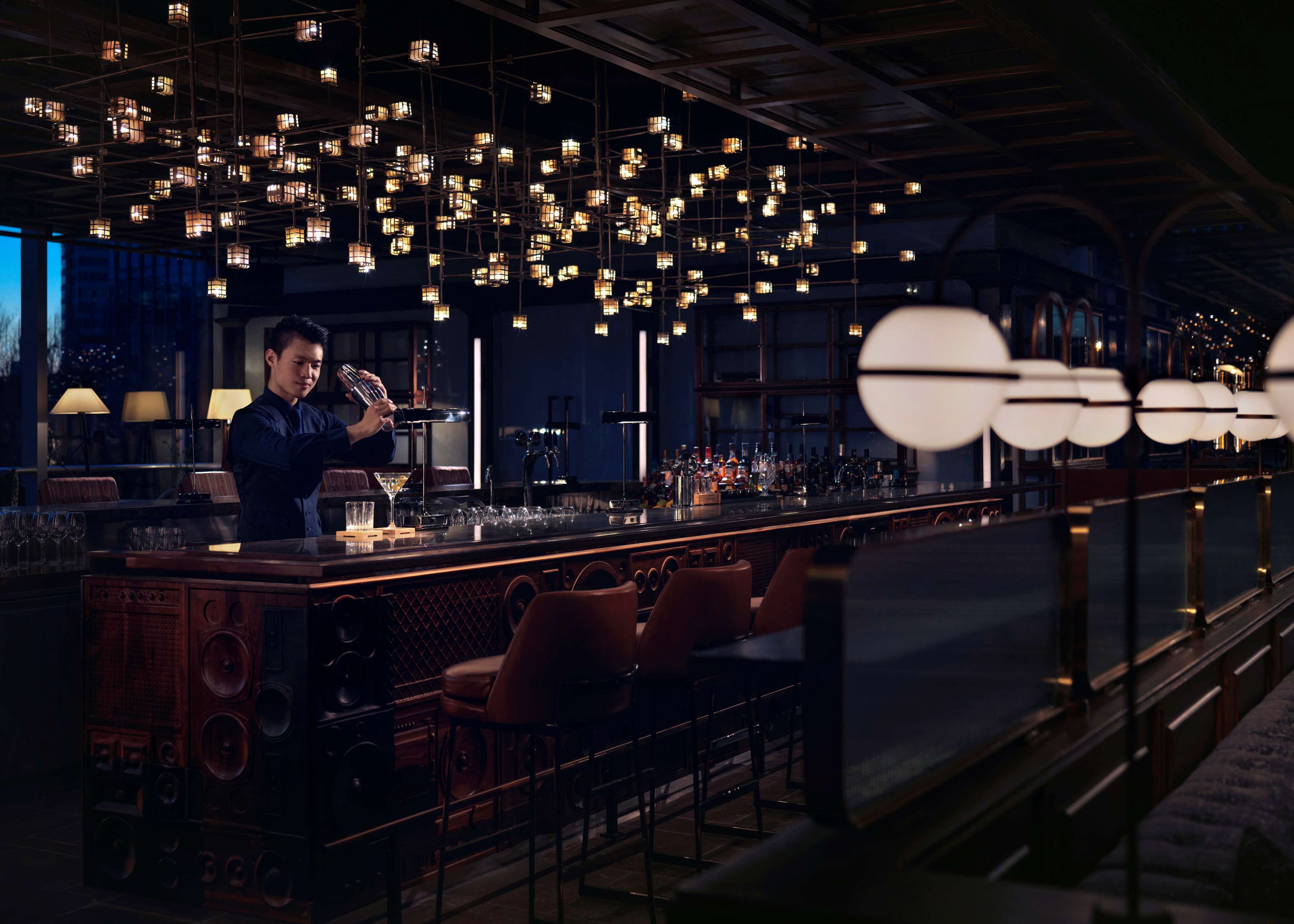 A bar area with a bartender preparing drinks, hanging lights, and a bar counter with glasses and bottles.