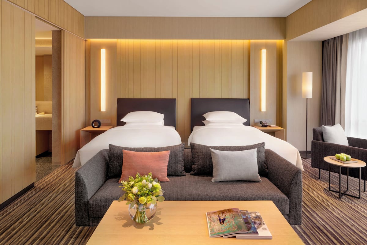 Una habitación de hotel con dos camas, un sofá, una mesa de centro con una revista y flores, y una lámpara.