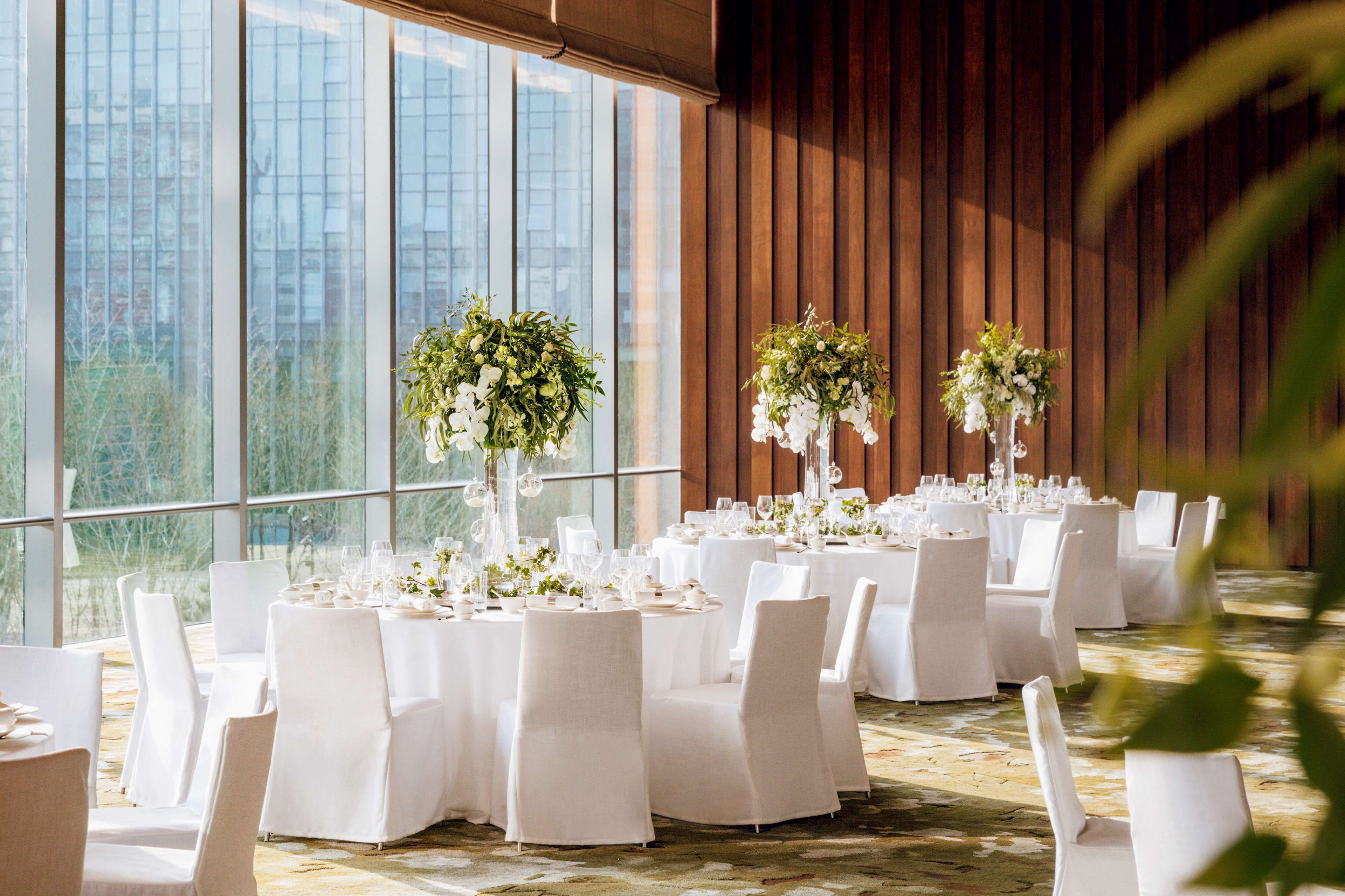 Un salón de banquetes con mesas redondas dispuestas para un evento formal, ventanas amplias y vistas panorámicas de la ciudad.
