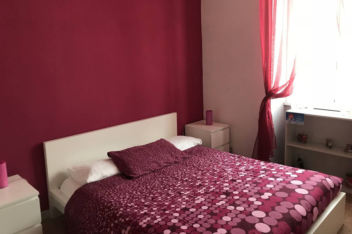 Un dormitorio con cama, mesitas de noche blancas, cómoda y ventana con cortinas.
