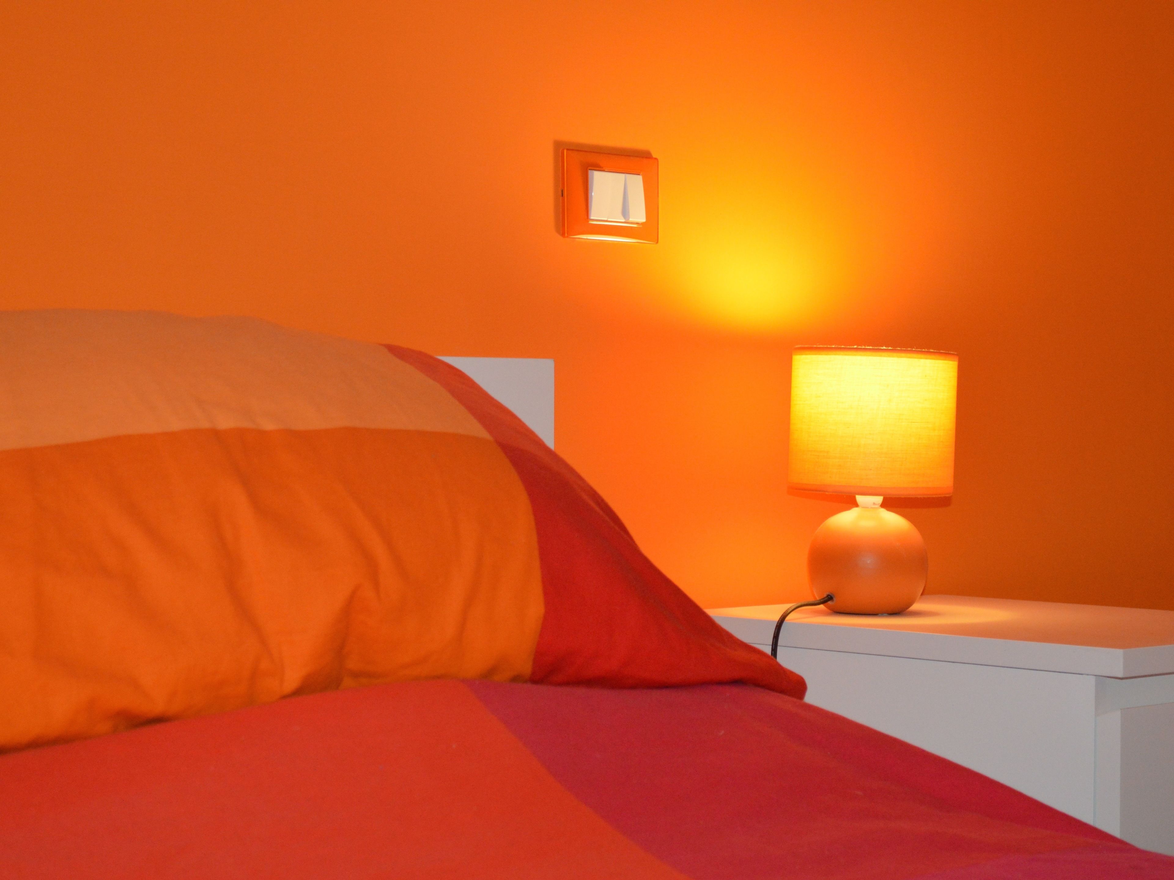 Uma cama com cobertor vermelho e laranja, uma mesa de cabeceira branca e um abajur com cúpula branca.