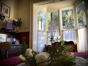 Quarto com uma janela grande, uma cadeira de madeira, uma mesa com um vaso de flores e um quadro na parede.