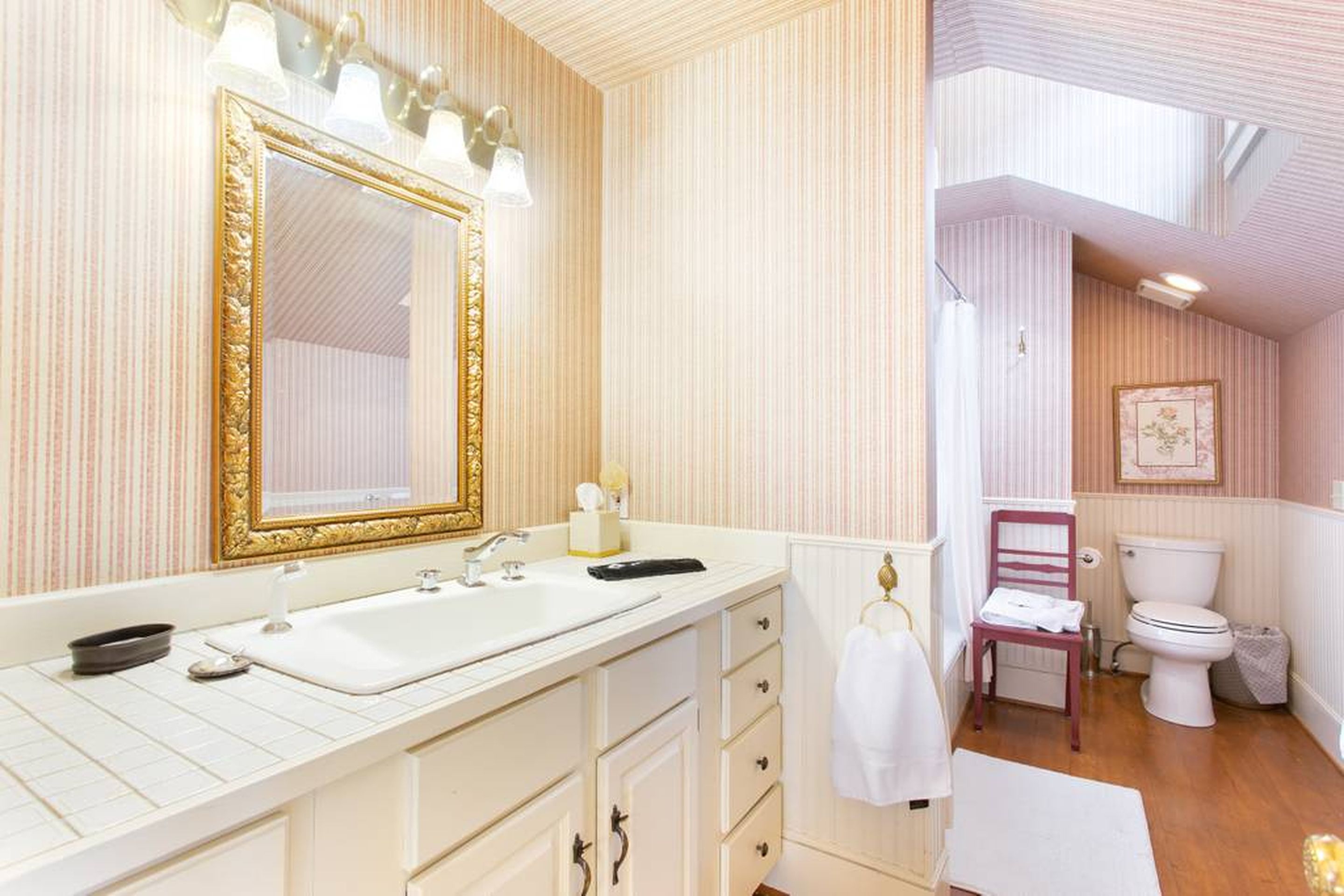 A bathroom with a large mirror, white sink, and vanity. There is a toilet and a chair in the adjacent room.