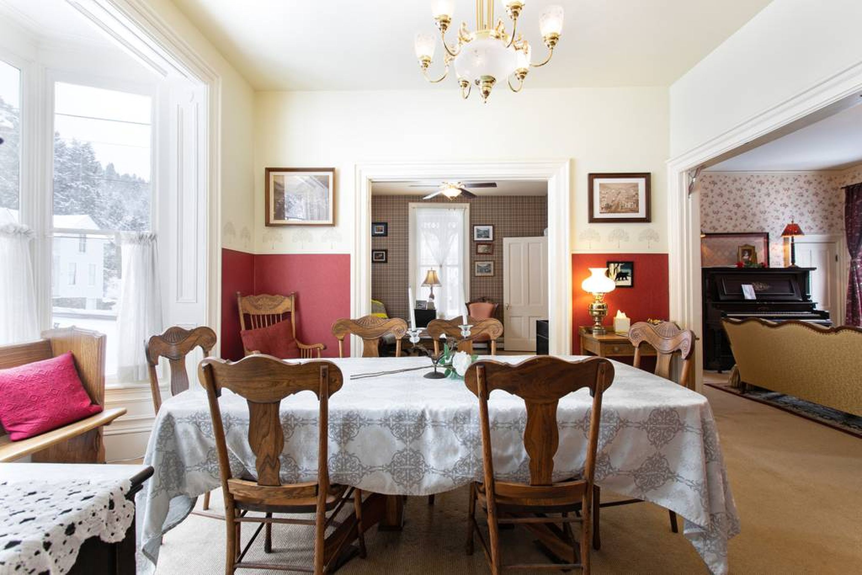 A dining room with a table set for four, wooden chairs, a chandelier, and a piano in the background.