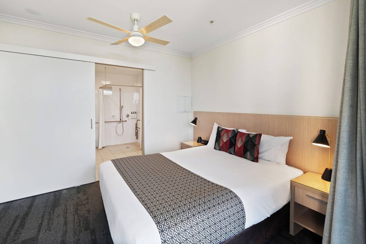 Habitación de hotel con cama, mesitas de noche, ventilador de techo, zona de ducha y armario.
