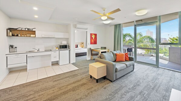 Une chambre d’hôtel moderne dotée d’une kitchenette, d’un canapé gris agrémenté de coussins orange, d’une petite table basse en bois et d’un balcon offrant une vue sur la ville et la verdure.