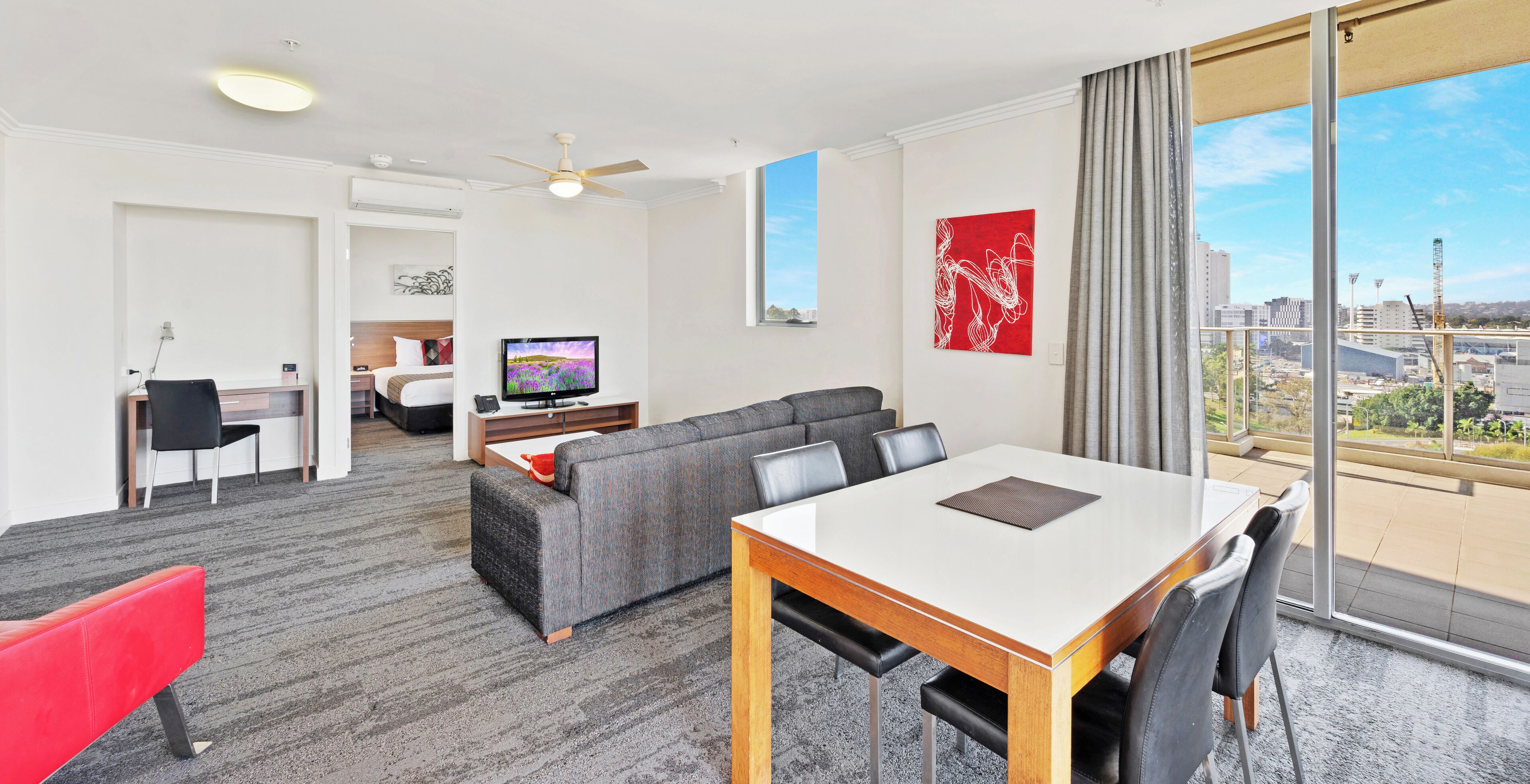 A modern hotel room with a dining area, a sofa, a TV, and a large window with a city view.