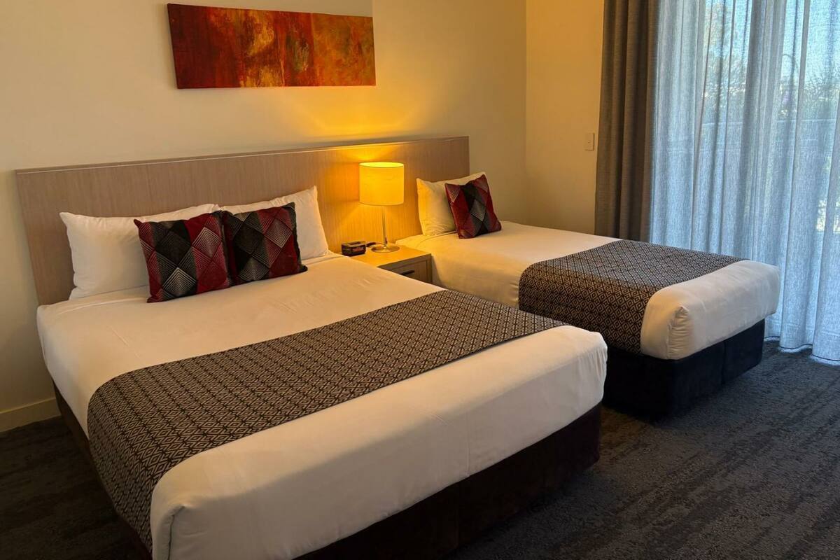 Habitación de hotel con dos camas, cada una con cubrecamas y almohadas estampadas, una lámpara de noche, un cuadro en la pared y vistas al exterior a través de cortinas translúcidas.