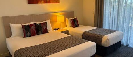 Habitación de hotel con dos camas, cada una con cubrecamas y almohadas estampadas, una lámpara de noche, un cuadro en la pared y vistas al exterior a través de cortinas translúcidas.