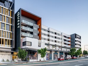 A modern multi-story hotel with a prominent "QUEST" sign, outdoor seating area, and a street view with parked cars.