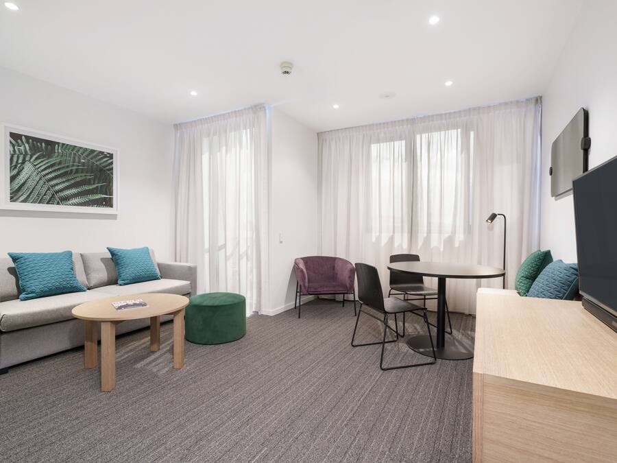 A modern hotel room with a grey sofa, a small round table, a green ottoman, a purple chair, and a TV on a wooden stand.