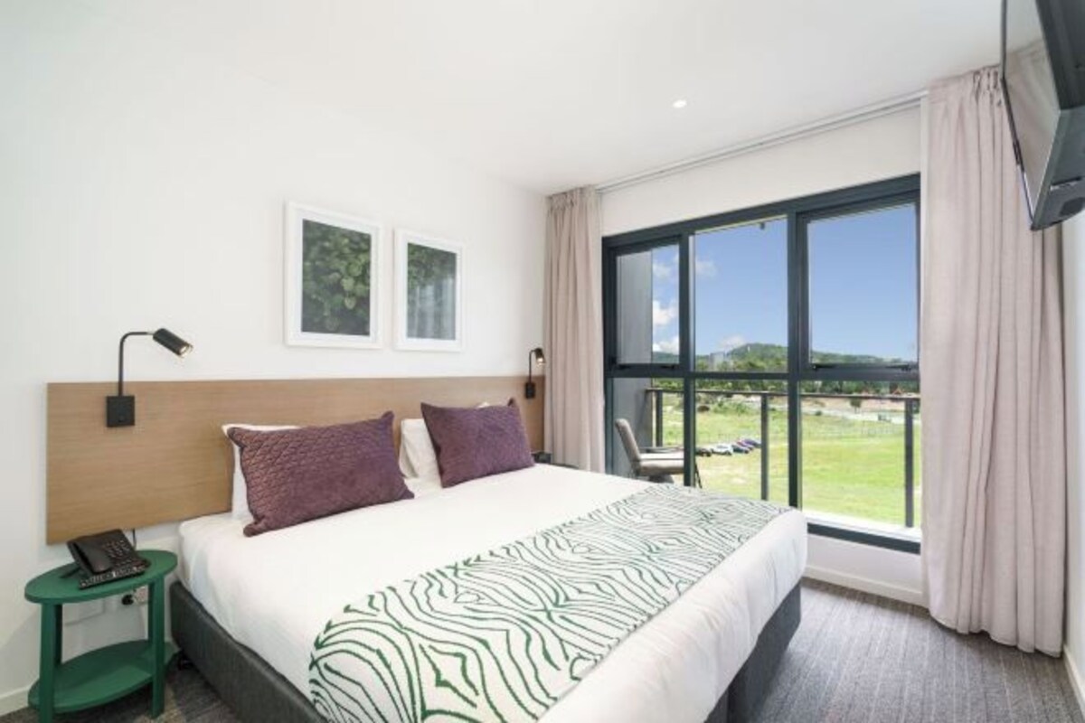 Habitación de hotel con una cama grande, una mesita de noche, un televisor y un ventanal con vistas al exterior.