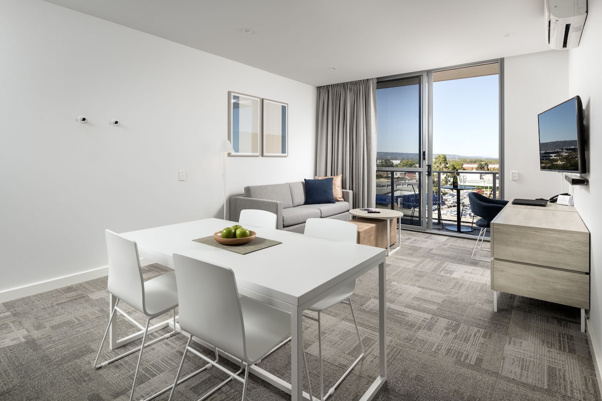 A modern hotel room with a dining area, a sofa, a TV, and a balcony with a city view.