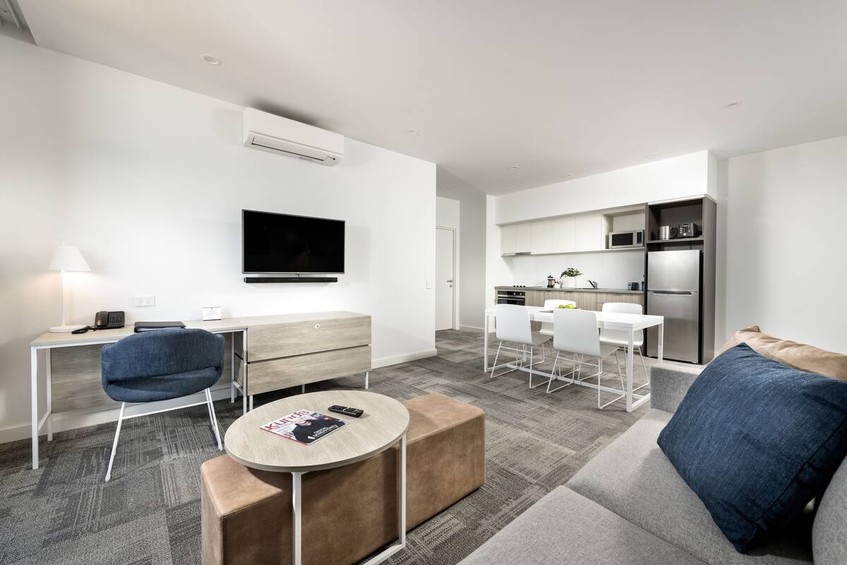 A modern, well-lit living room with a flat-screen TV, a dining area with white chairs, and a kitchen with stainless steel appliances.