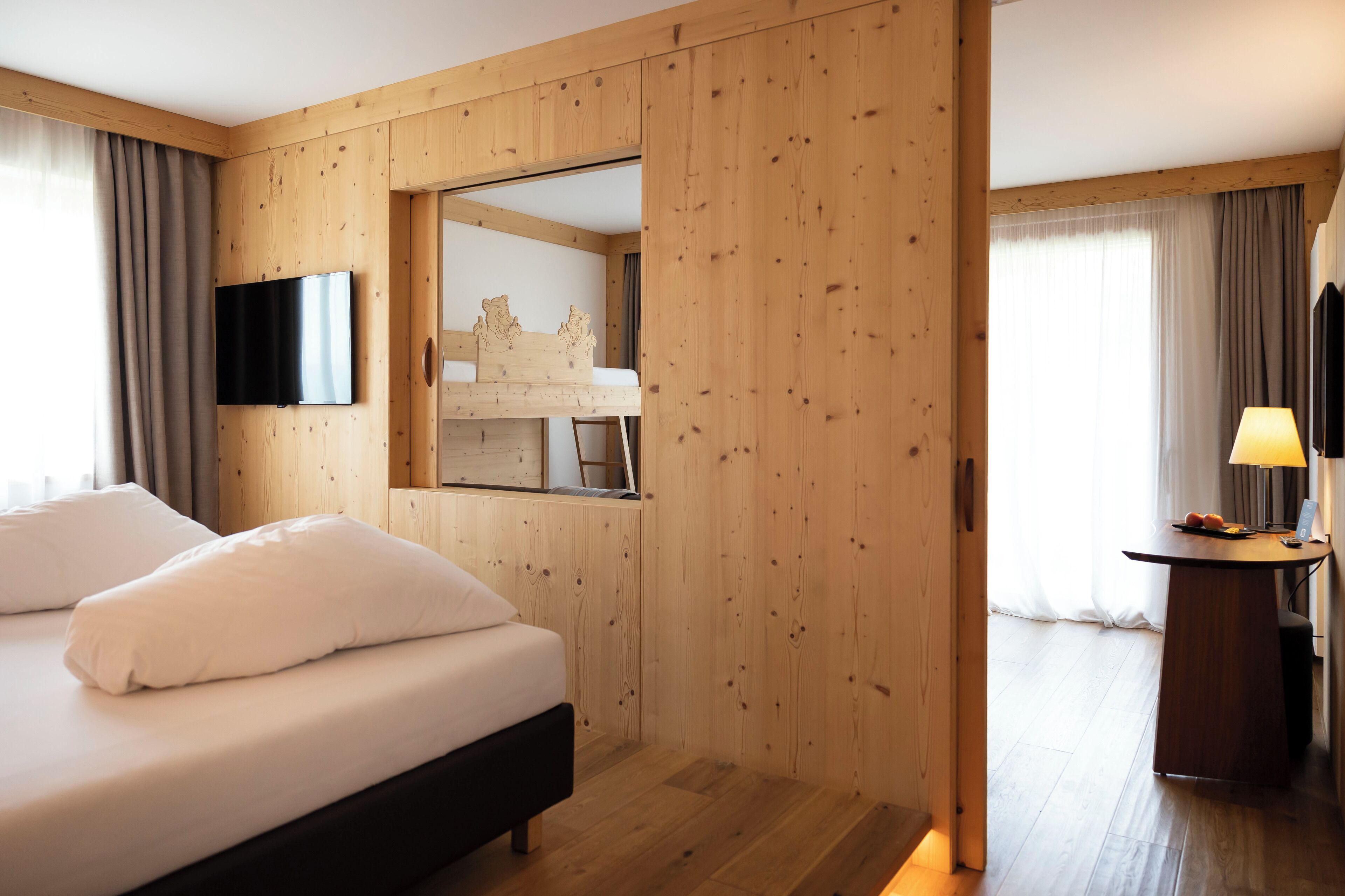 A hotel room with a wooden paneled wall, a bed with white bedding, a television mounted on the wall, and a desk with a lamp.
