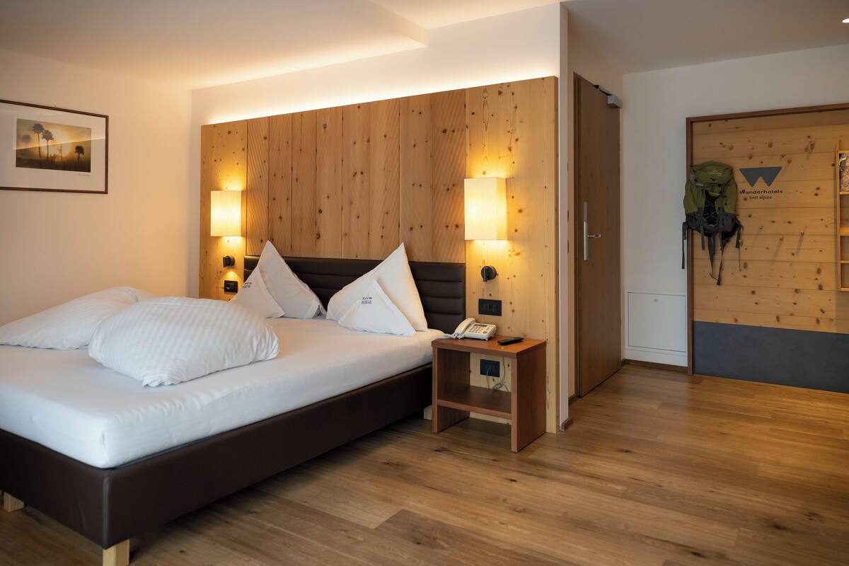 A modern hotel room with a wooden headboard, a bed with white linens, bedside lamps, a small wooden nightstand, and a backpack hanging on the wall.