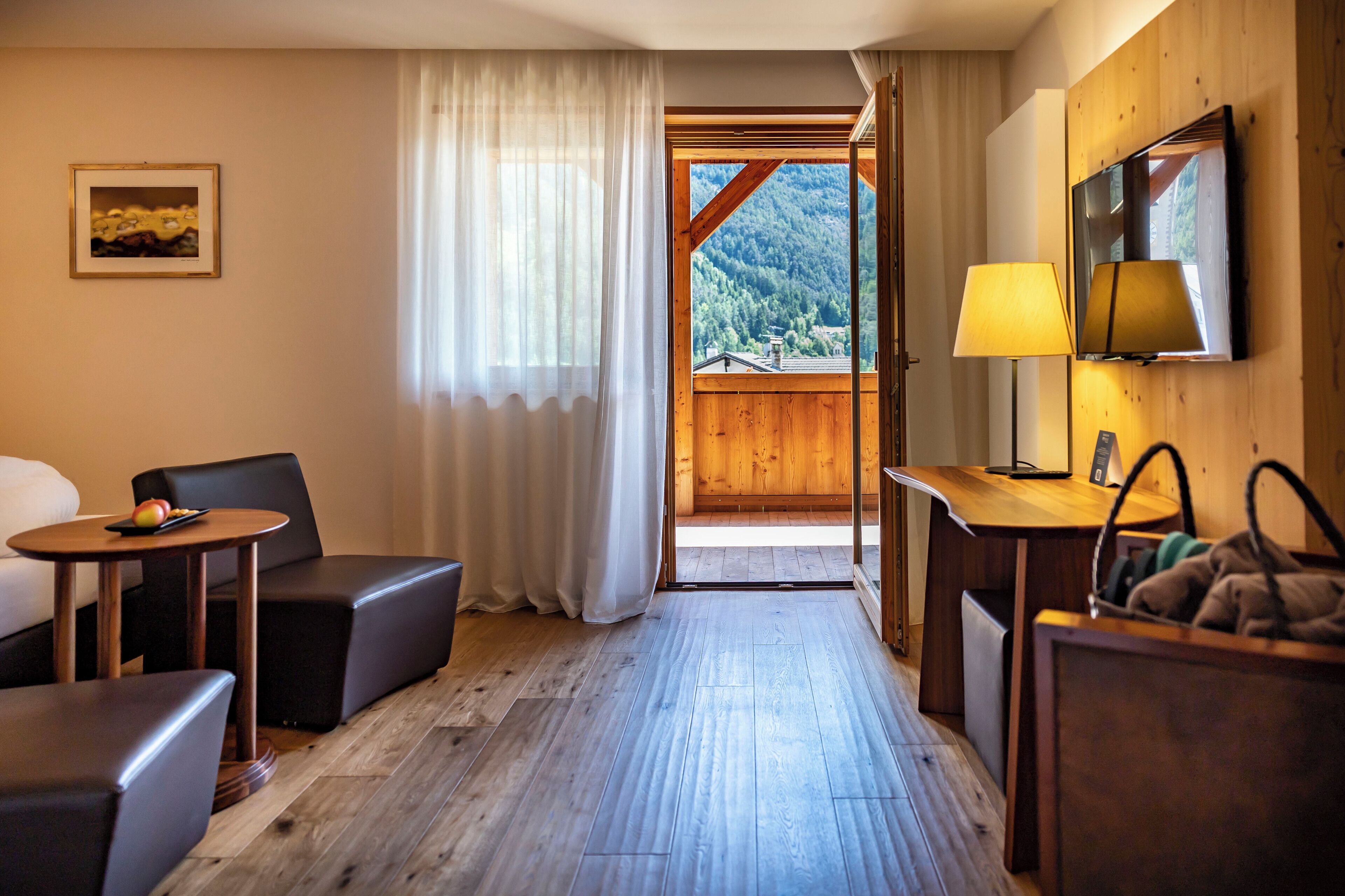 A hotel room with a wooden floor, a bed, a desk with a lamp, a TV, and a view of a mountainous landscape through a window.