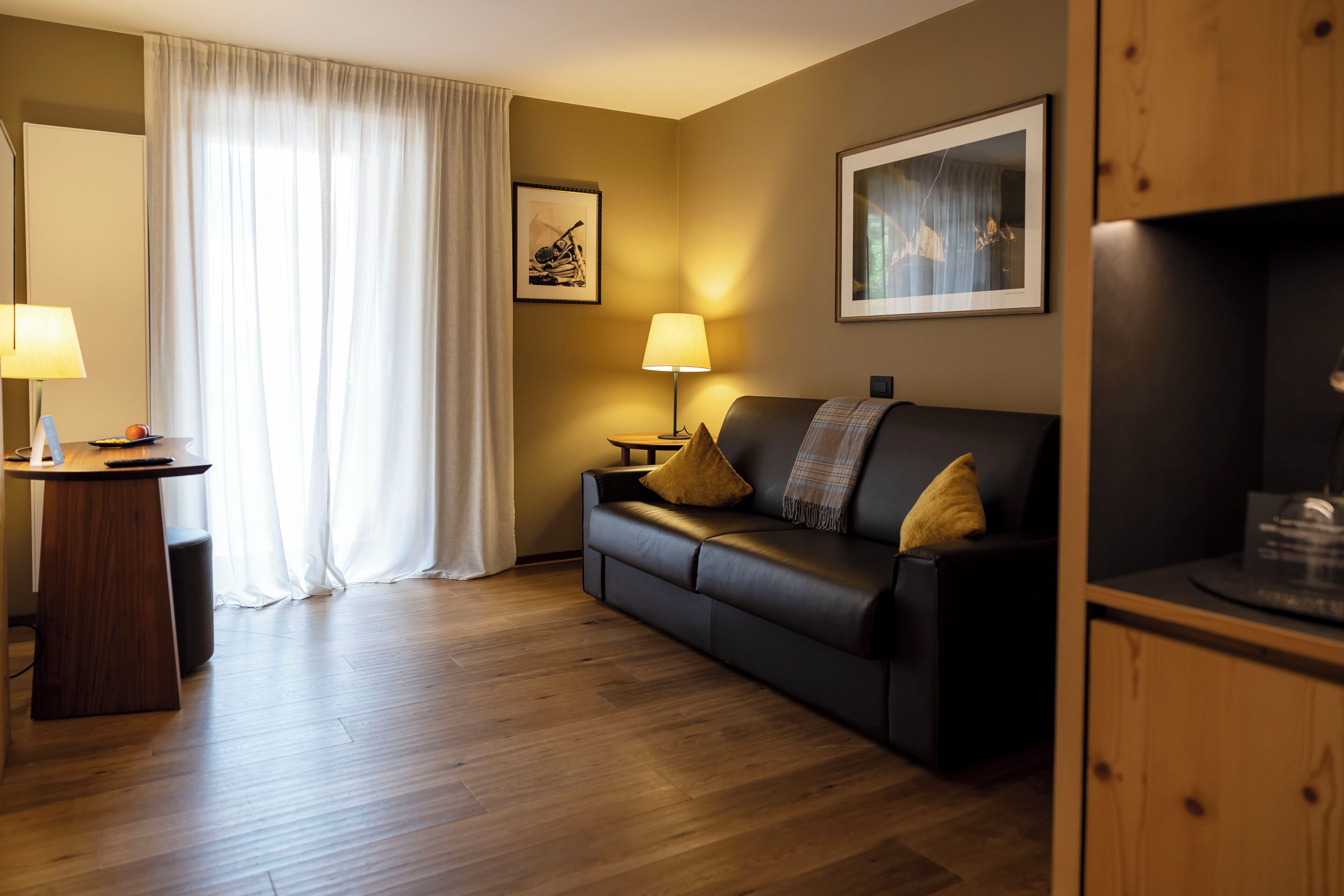 Ein modernes Hotelzimmer mit Holzboden, einer schwarzen Ledersofa, einem Holztischchen mit Lampe und einem großen Fenster mit durchsichtigen Vorhängen.