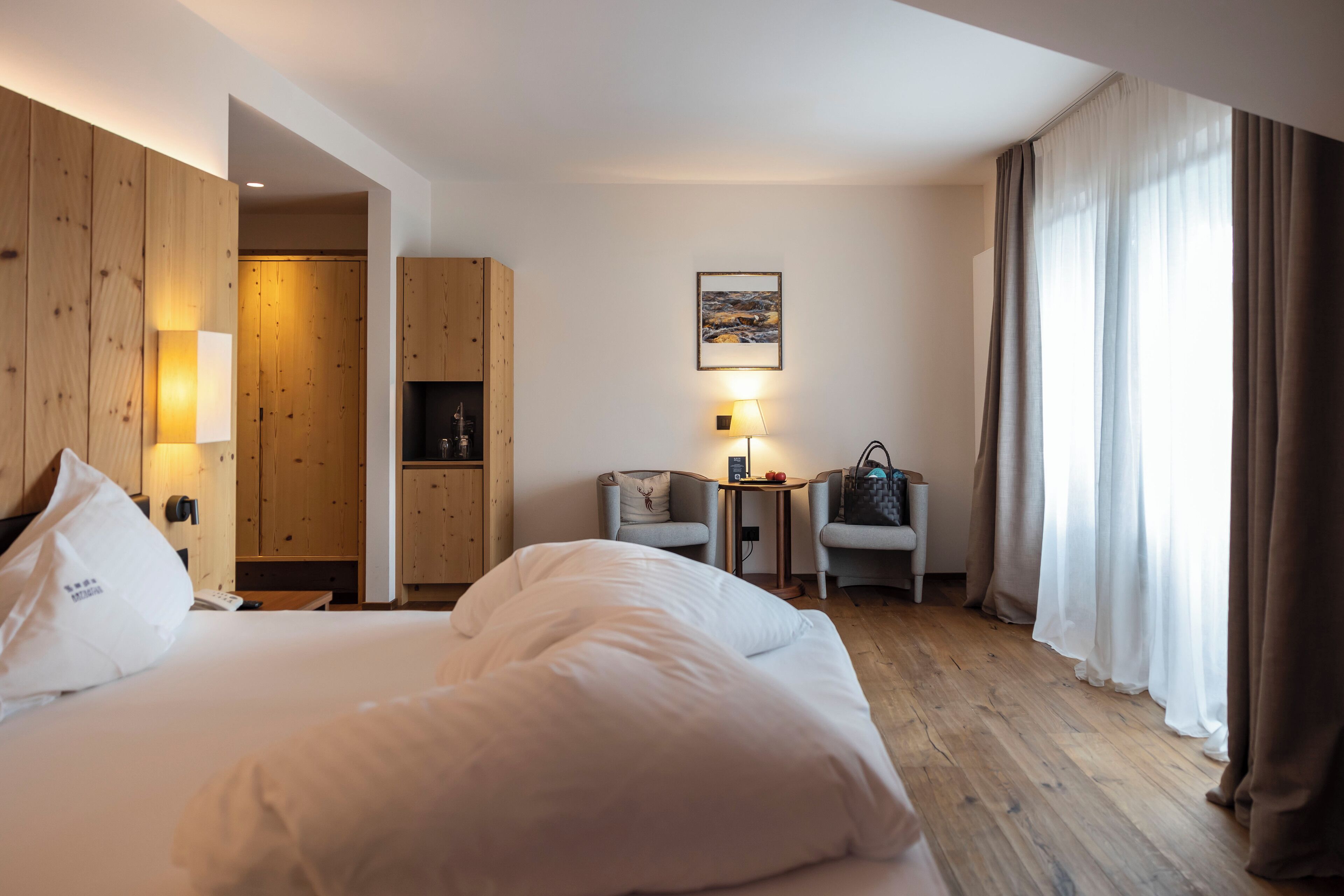 A hotel room with a large bed, wooden panel walls, a small table with a lamp, and a view through sheer curtains.