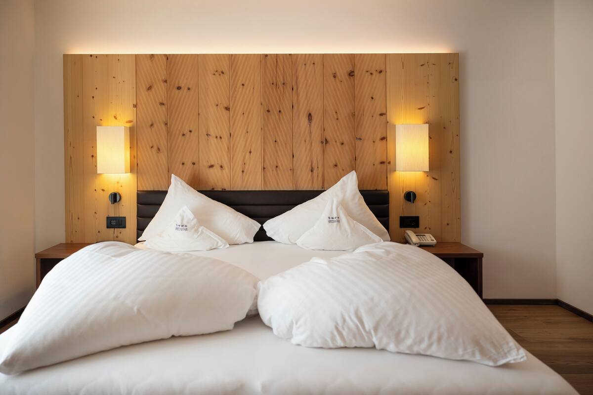 A double bed with white linens and pillows, wooden headboard, bedside lamps, and a nightstand with a phone.