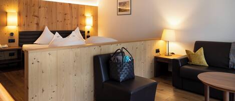 A hotel room with a wooden headboard, a bed with pillows, a black ottoman, a sofa, a small table, and a nightstand with a lamp.