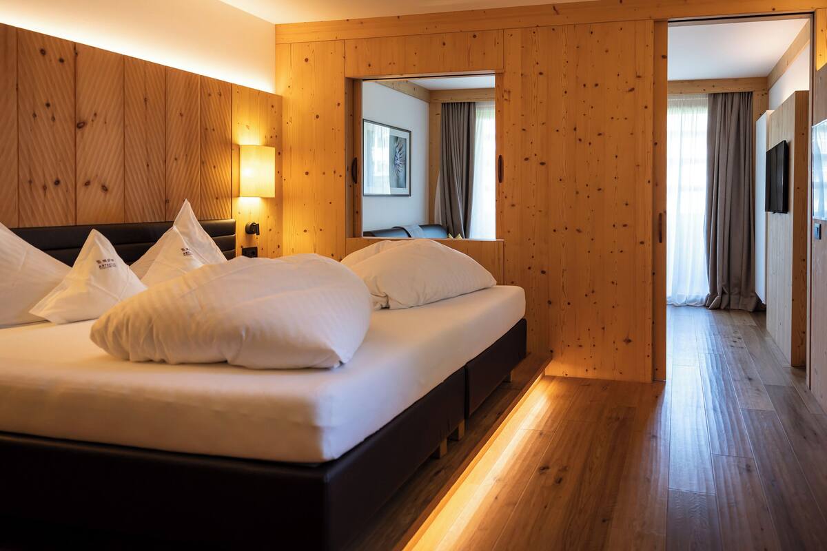 A hotel room with a wooden wall, a bed with white bedding, a bedside table with a lamp, and a view through an open door.