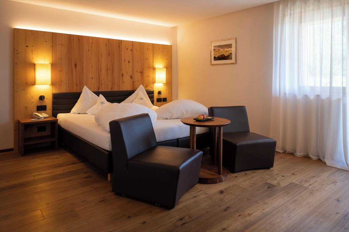 A hotel room with a wooden headboard, a bed with white linens, two black armchairs, a small round table with fruit, and a nightstand with a phone.