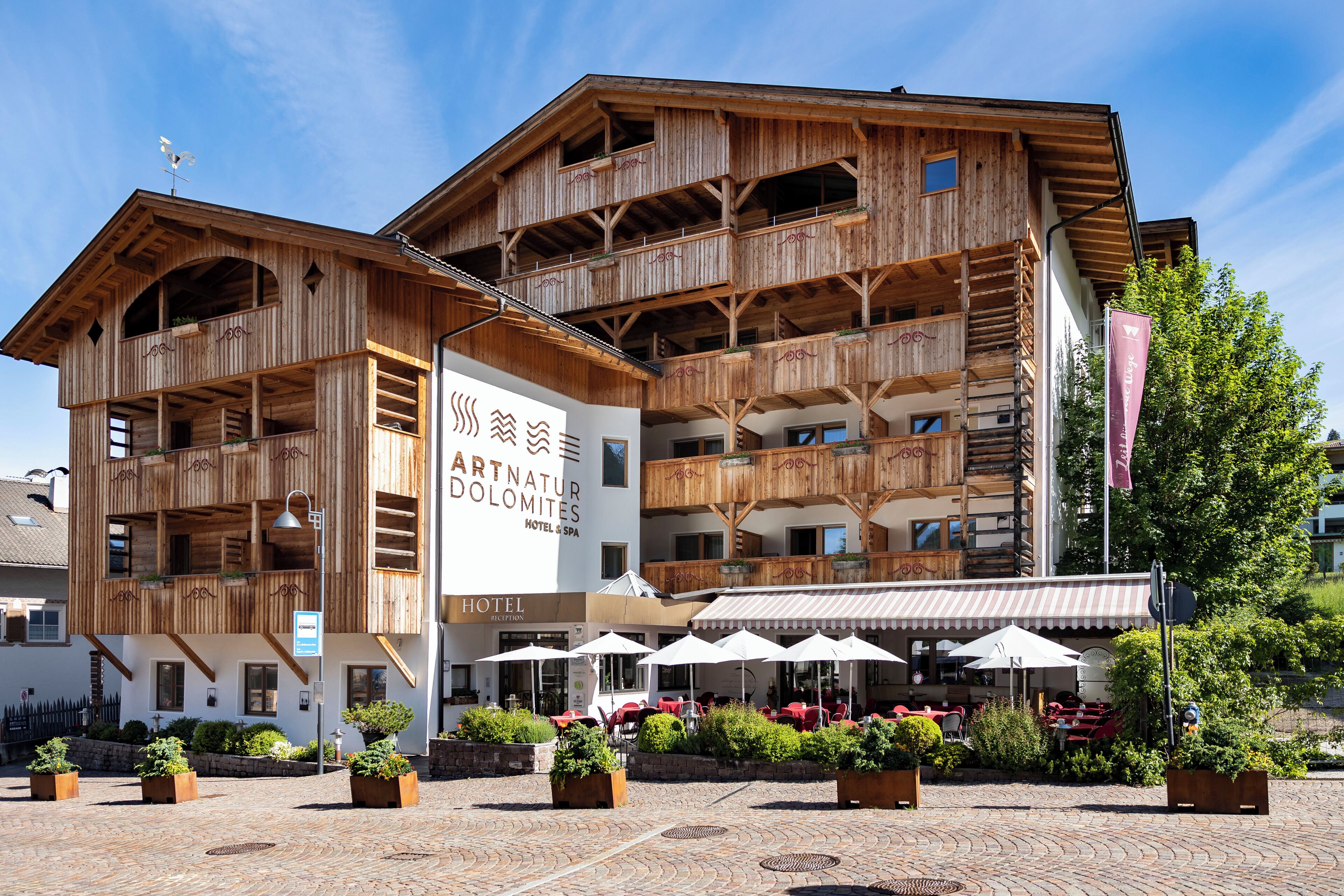 En træbygning med balkoner, et skilt med 'Art Natur Dolomites Hotel & Spa', udendørs siddepladser med parasoller og plantekasser.
