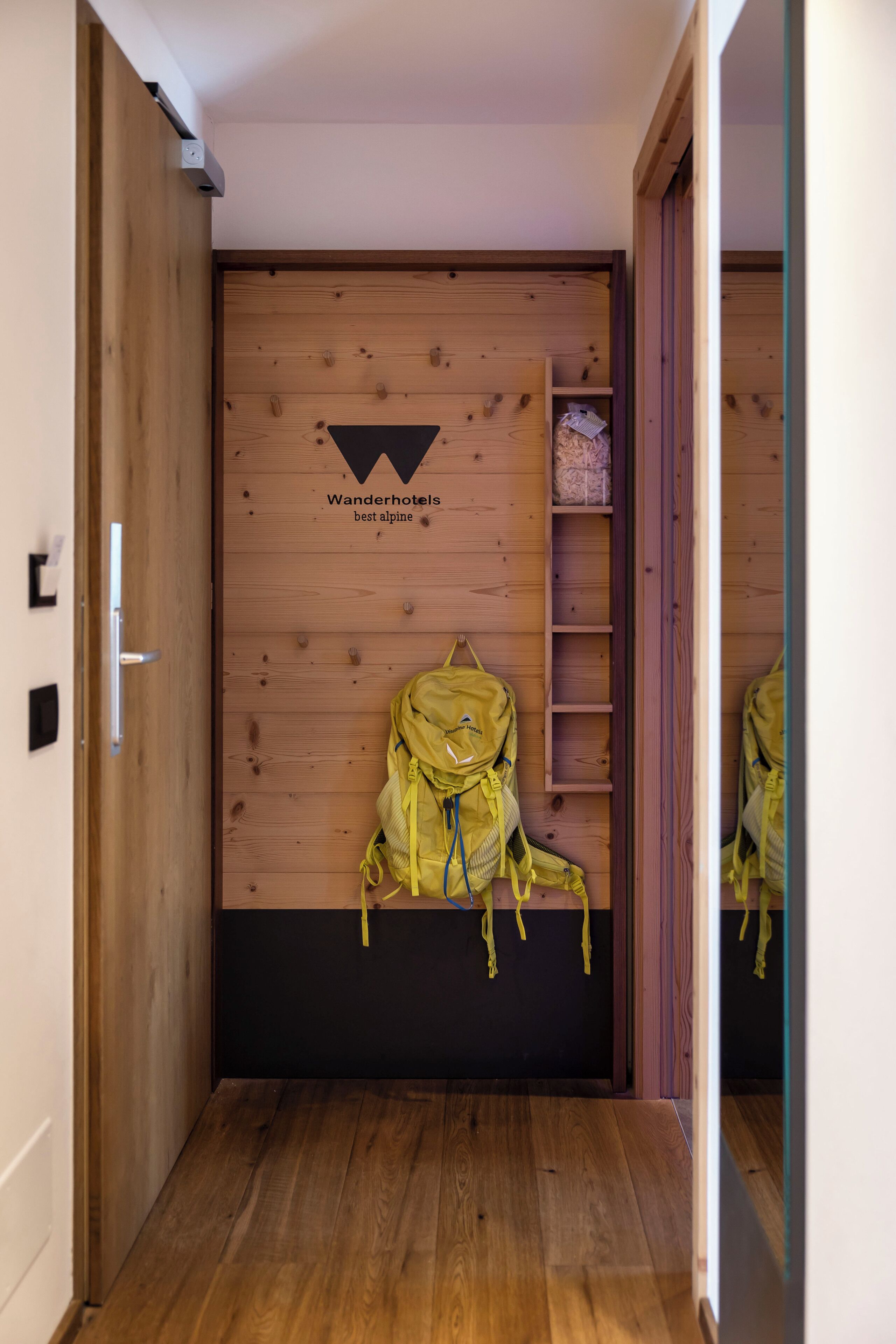 Ein Flur mit Holzwänden, ein gelber Rucksack hängt an der Wand, und ein Schild mit der Aufschrift 'Wanderhotels best alpine'.