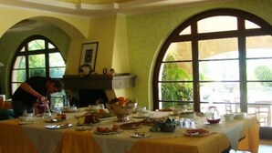 Uma sala de jantar com mesa posta para refeição, alguém servindo comida e grandes janelas arqueadas com vegetação visível lá fora.