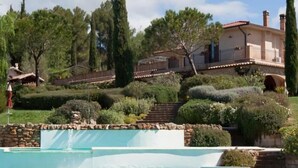 Uma casa grande com piscina, cercada por jardins bem cuidados e árvores.