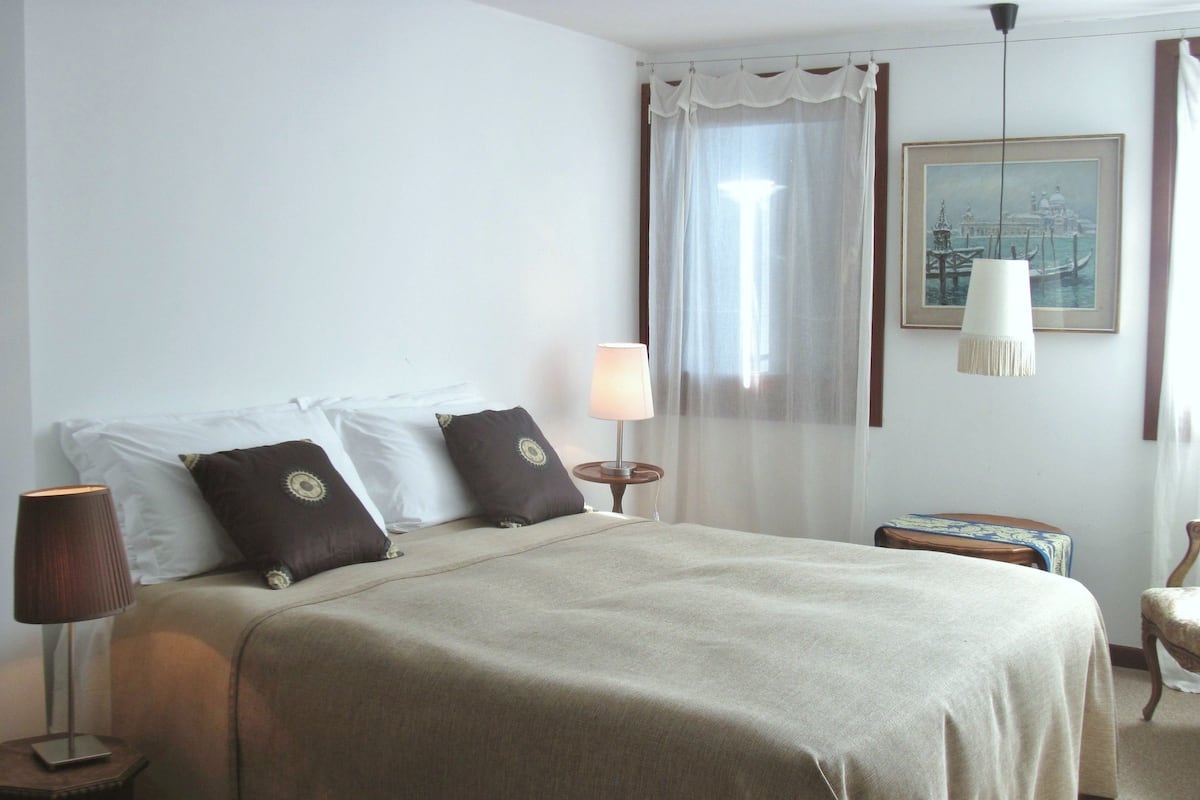 A hotel room with a bed, a bedside table, a lamp, a chair, and a painting on the wall.