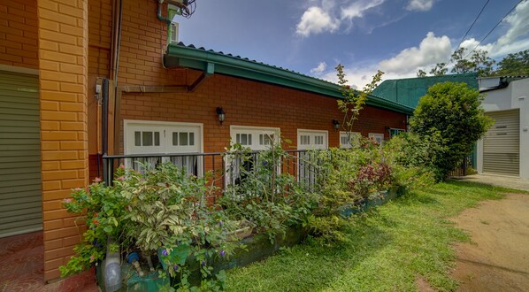 A brick building with two white-framed windows, a green roof, and a garden with various plants and flowers.