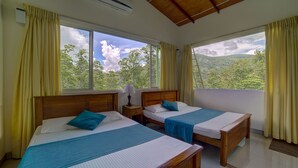 A room with two wooden beds, each with a blue bedspread and pillows, a nightstand with a lamp, and large windows offering a view of a forested landscape. - Hanthana Holiday Rooms (Kandy)