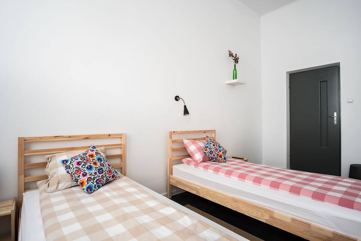 Quarto com duas camas de solteiro, uma pequena escrivaninha, uma cadeira, uma luminária e uma porta.