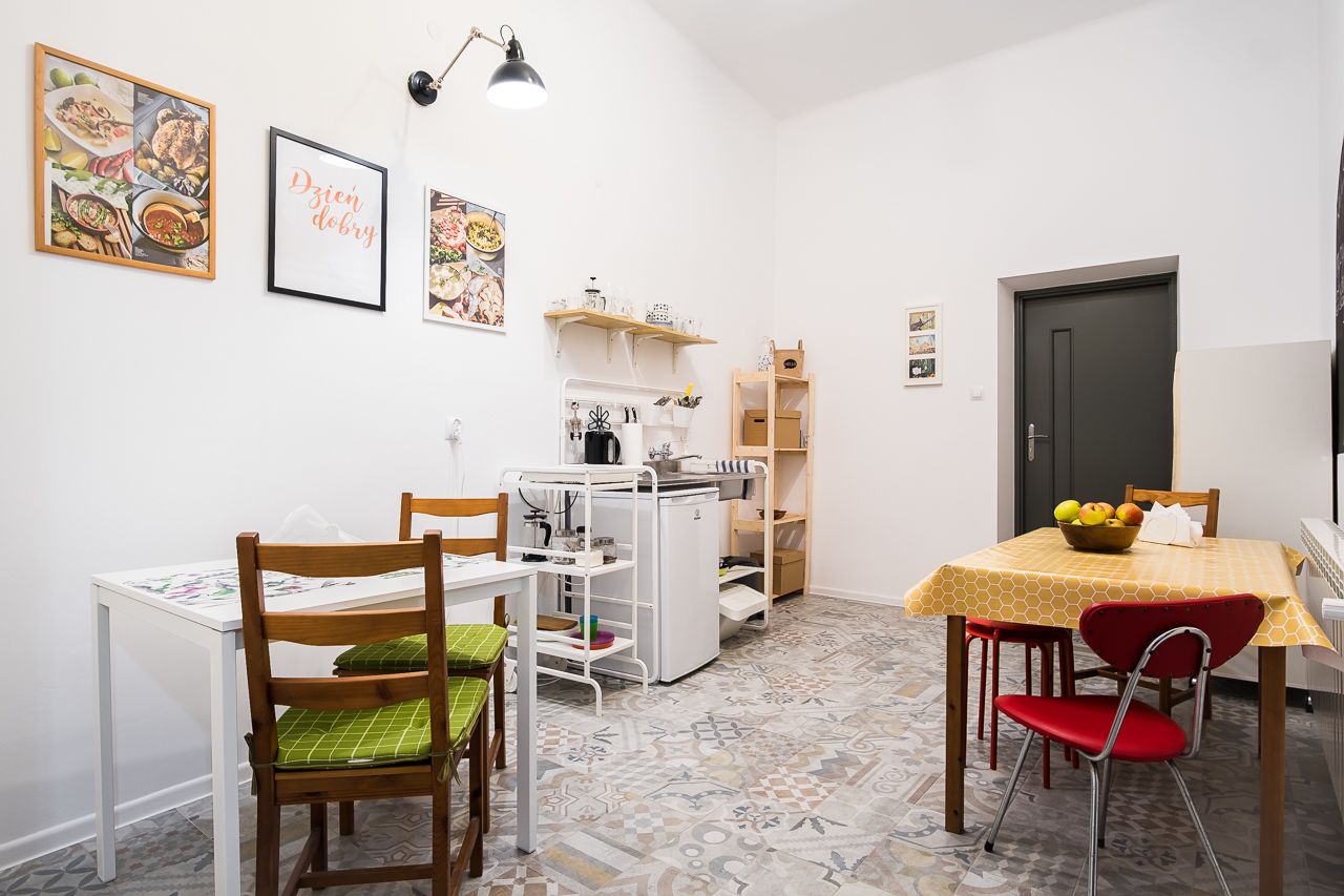 Una cucina compatta con un frigorifero bianco, una piccola isola e un tavolo da pranzo con sedie. Alle pareti ci sono quadri incorniciati e una porta a destra.