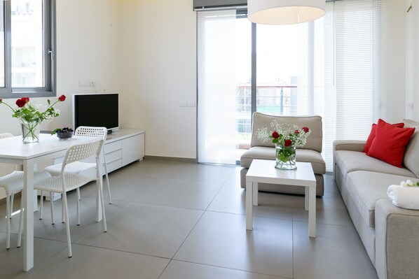 A modern living room with a white sofa, a dining table with chairs, a TV, and a large window with blinds.